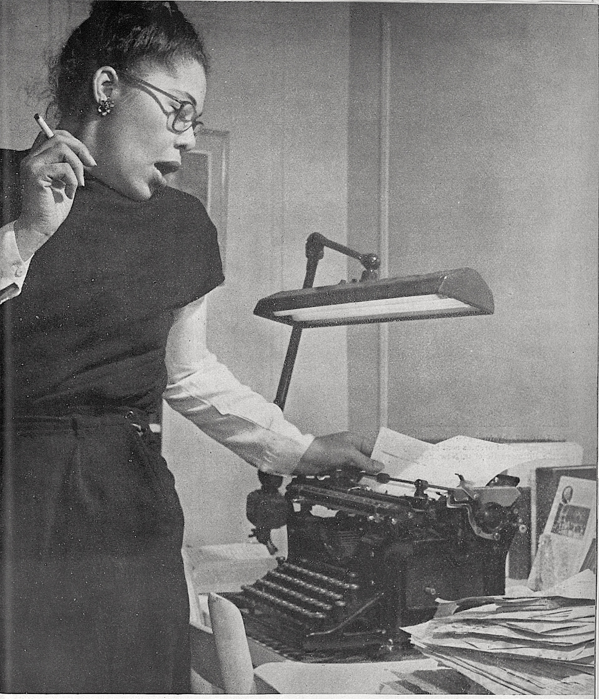 Ann Petry standing holding a cigarette with her right hand and holding paper in a typewriter with her left. 