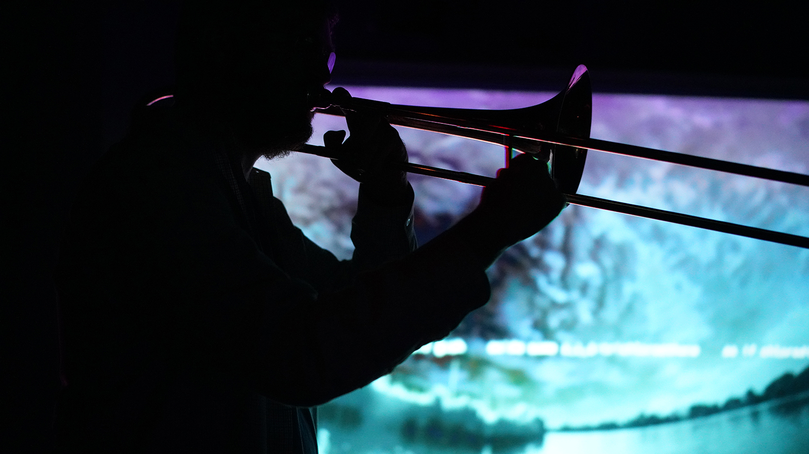 A trombone player plays against a video screen lit with indecipherable text and images