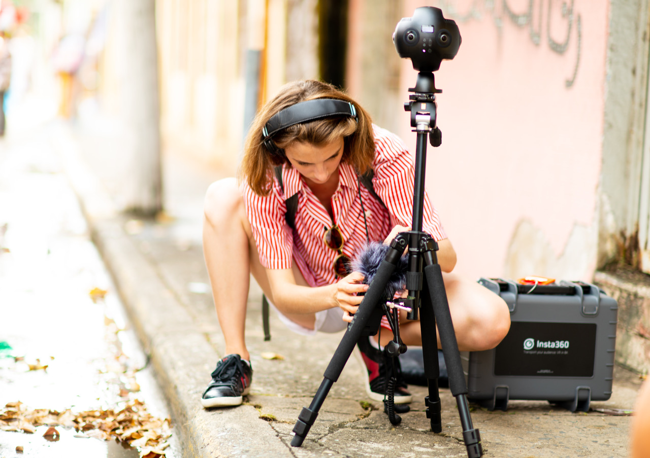 A person wearing headphones crouches down to set up a camera on the sidewalk