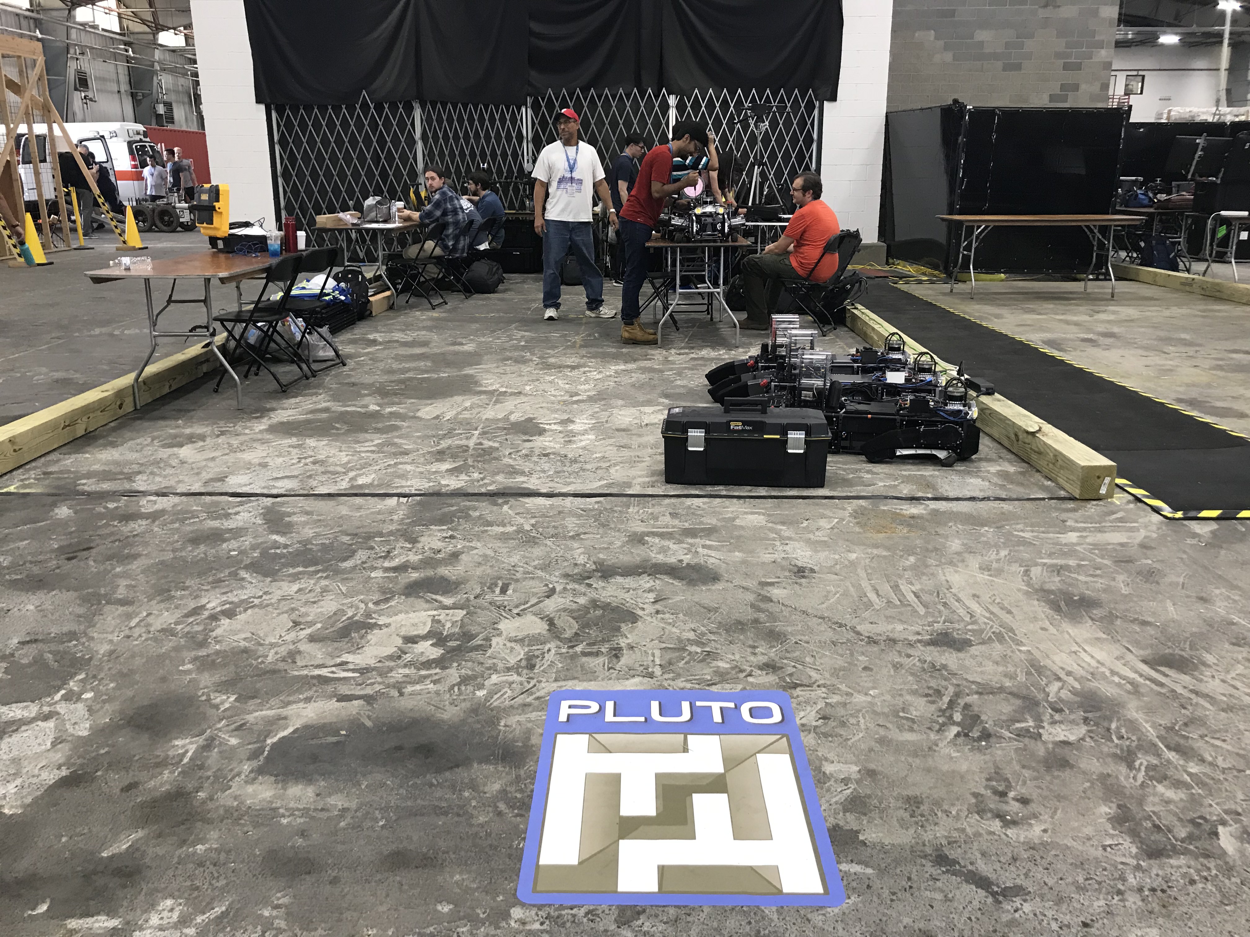 the PLUTO logo on a concrete floor with a team of students in the background sitting on chairs at tables and the folded up dog robots in front of the people