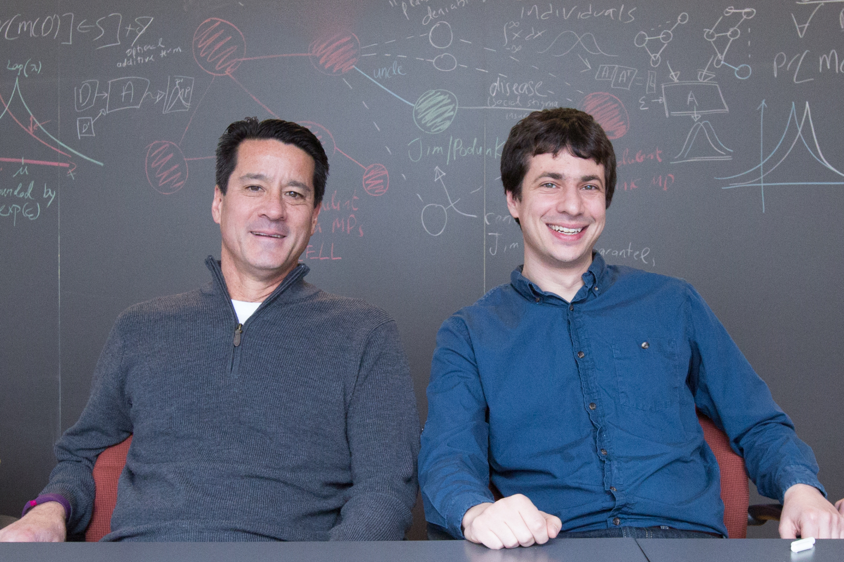 michael kearns and aaron roth sitting at a table in front of a chalk board