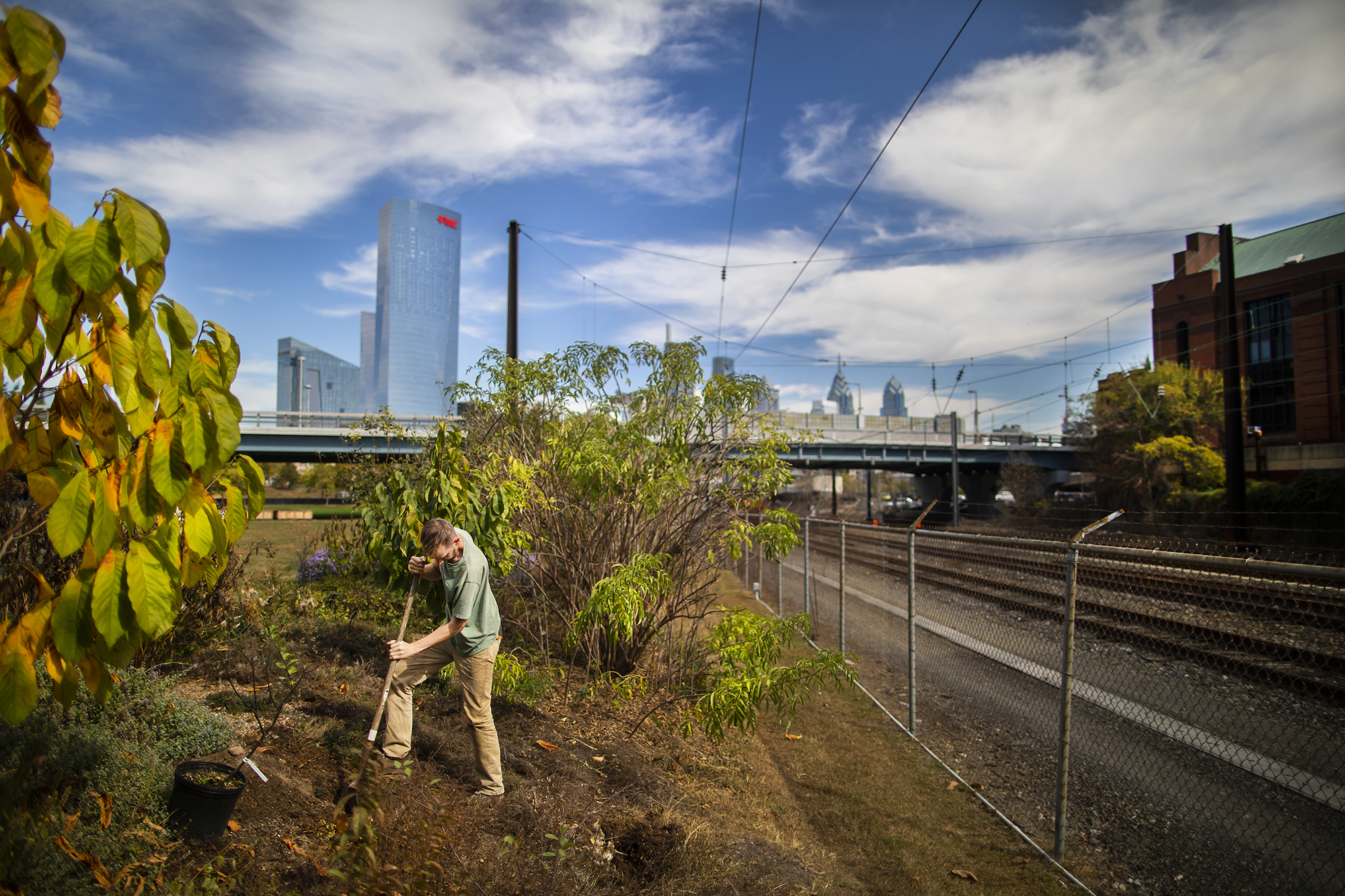 Person digs with shovel in garden bed next to railroad tracks, with the Philadelphia skyline in the background