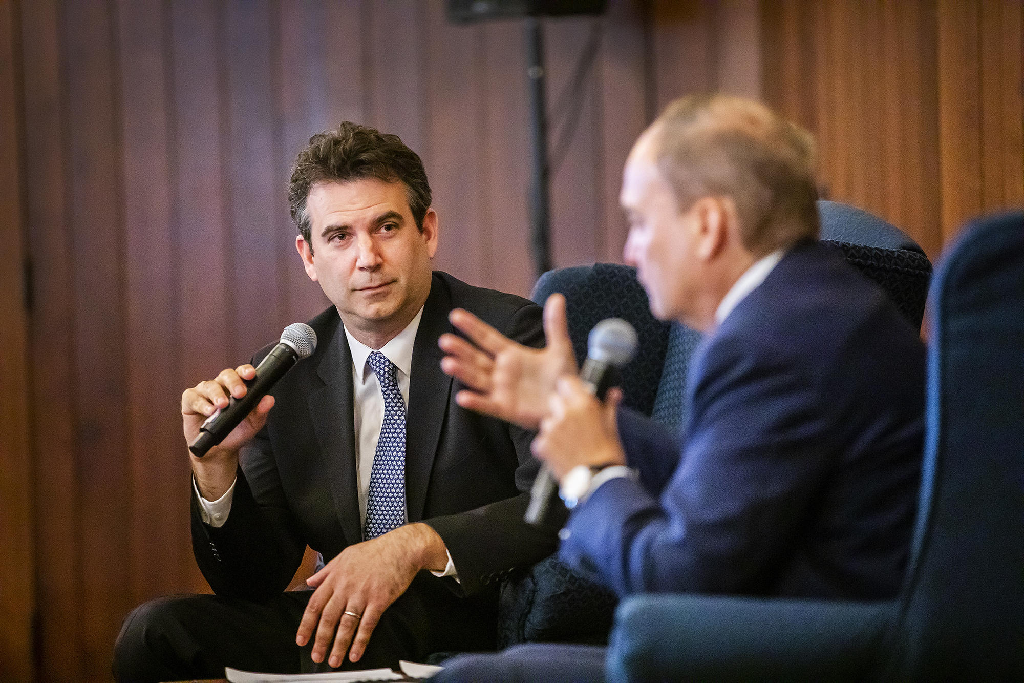 Person holding microphone sitting in blue wing-backed chair looks at another person sitting in a blue wing-backed chair who is speaking into a microphone