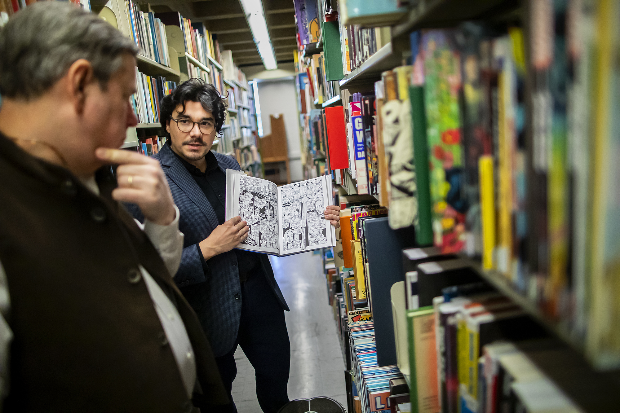 J.C. Cloutier standing by shelves full of books holding open a book with drawings of comics.