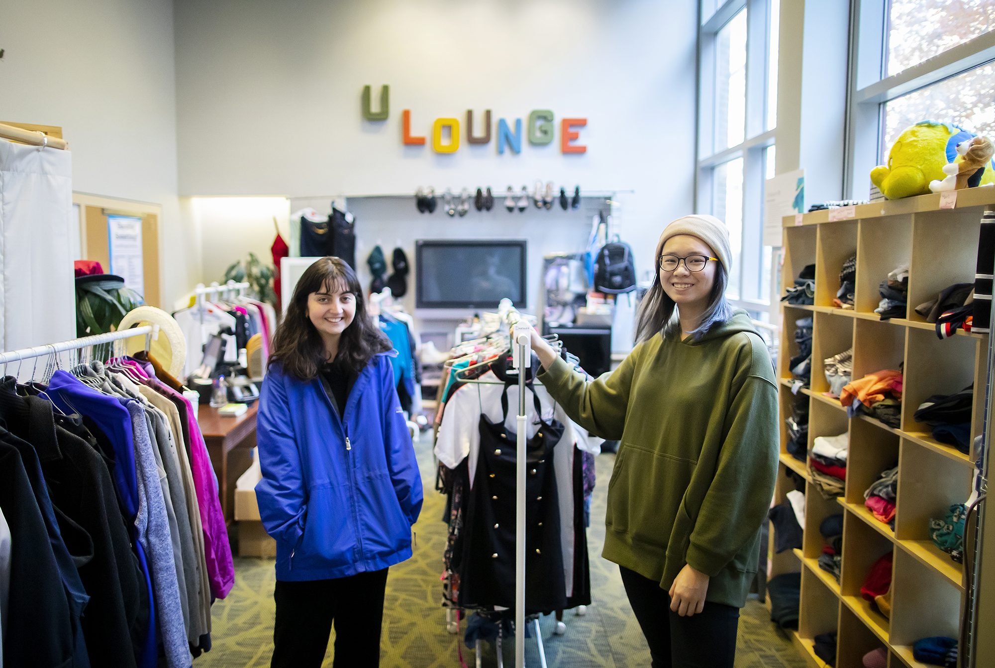 Two people standing bewteen two clothing racks in a crowded thrift shop space. On the back wall, the word "U Lounge" hangs in brightly colored block letters.