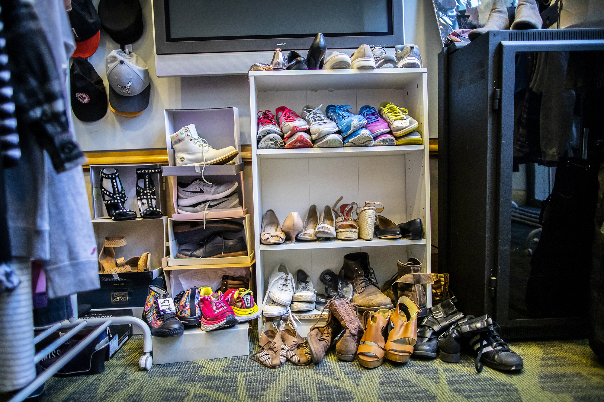 Many pairs of shoes in different styles, on three shelves and spilling onto the floor. A television is above them, and several hanging hats sit to the left. Clothes in the foreground are blurry.