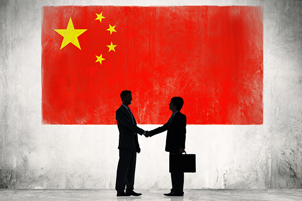 Two figures in business suits shake hands under a large Chinese flag.