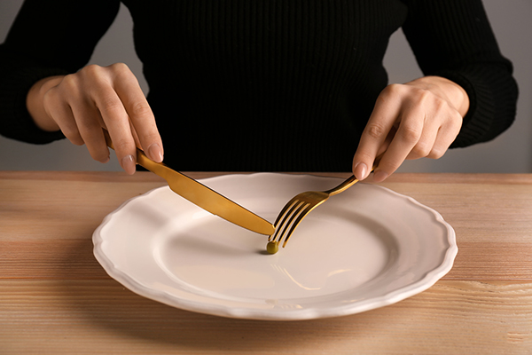 closeup of a person's hands cutting a single pea with a fork and knife on a dinner plate