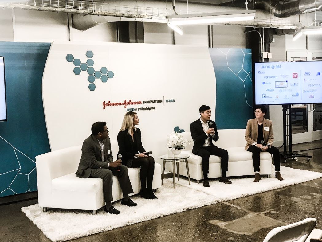 four people sitting on two couches holding microphones, one person is speaking to an audience out of view, they are next to a large screen dotted with company logos and infront of a large banner that reads Johnson and Johnson Innovation JLABS, JPOD at Philadelphia