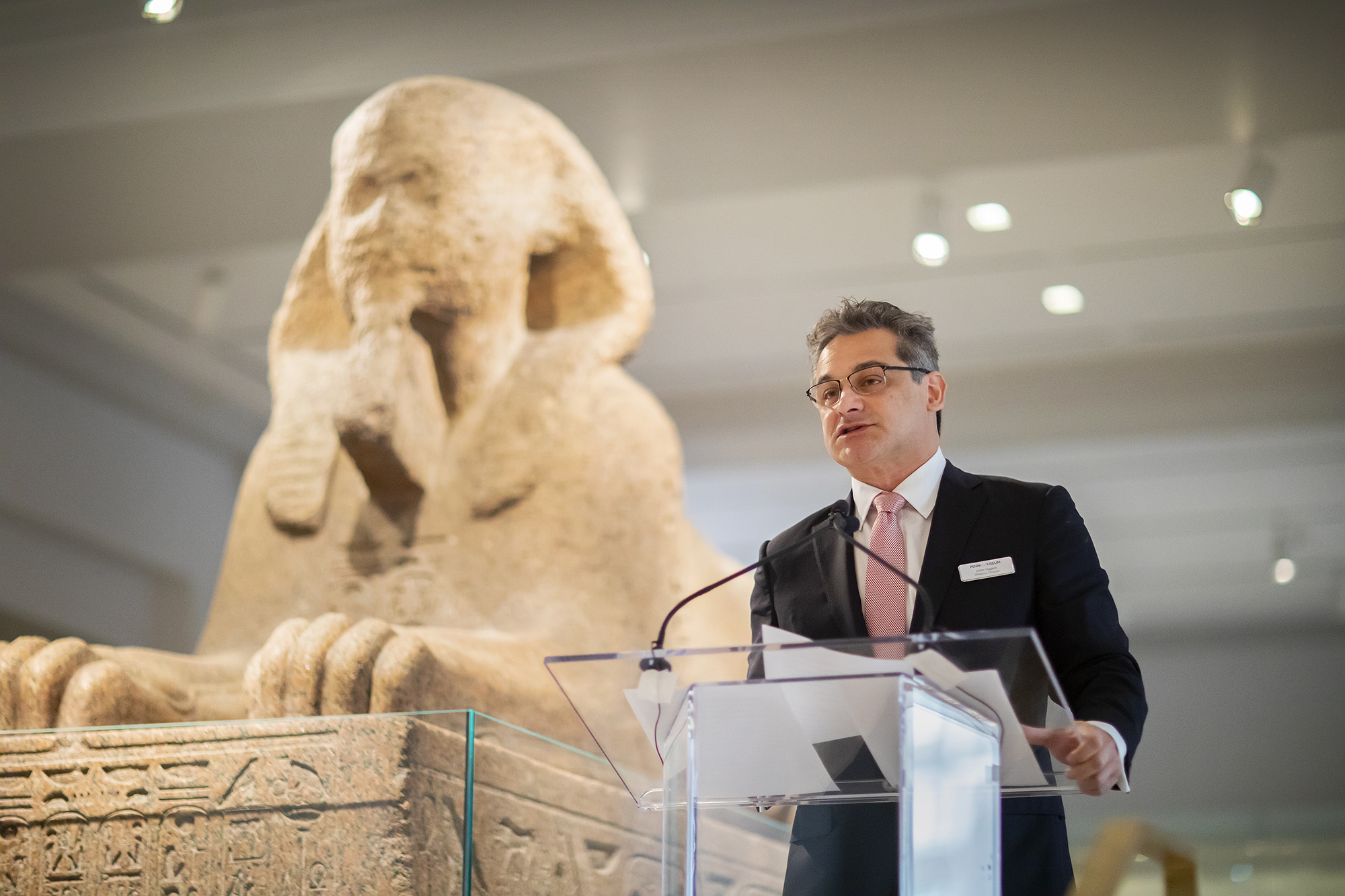 penn museum director julian siggers speaking at a podium with the sphinx behind him