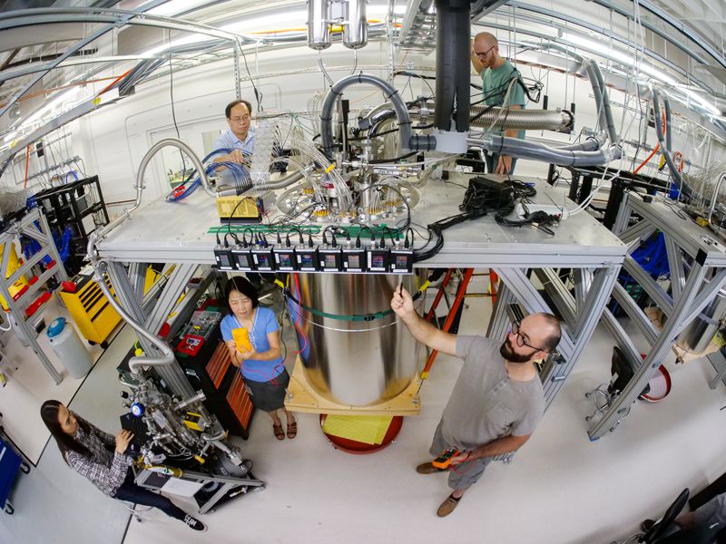 five people standing around a large metal vessel with wires coming out of the top