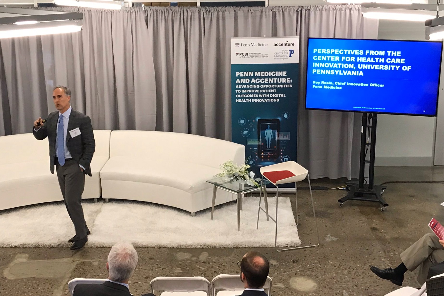 a person in front of a white couch and gray curtains and a TV screen that says perspectives from the center for health care innovation, university of pennsylvania giving a presentation to an audience