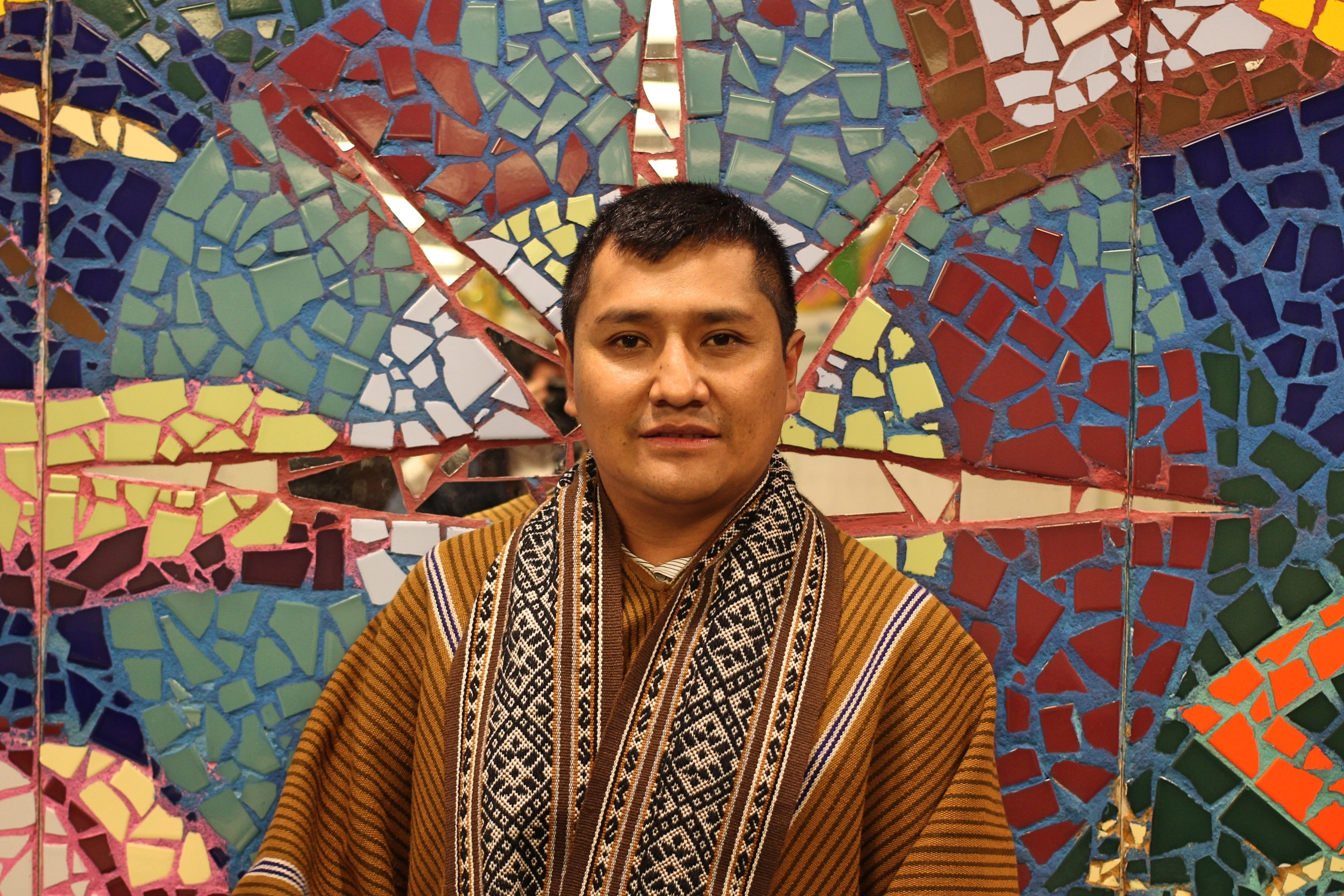 Man wearing traditional Andean clothing stands in front of mosaic