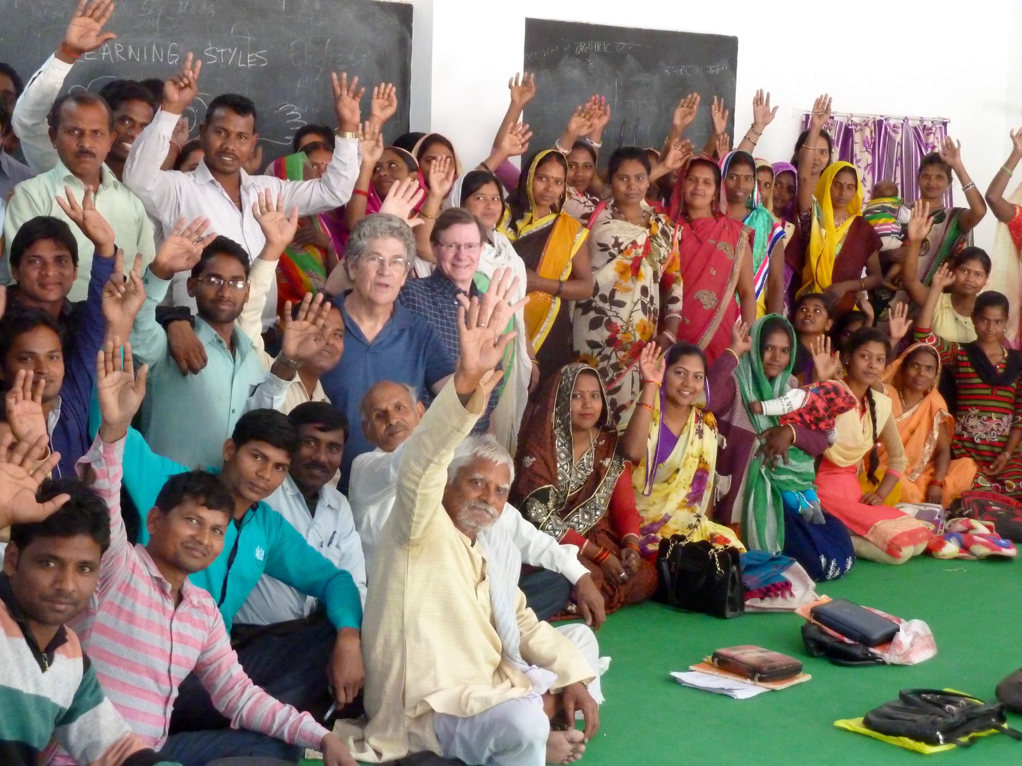 Classroom full of people seated and standing, many wearing saris, each raising a hand.