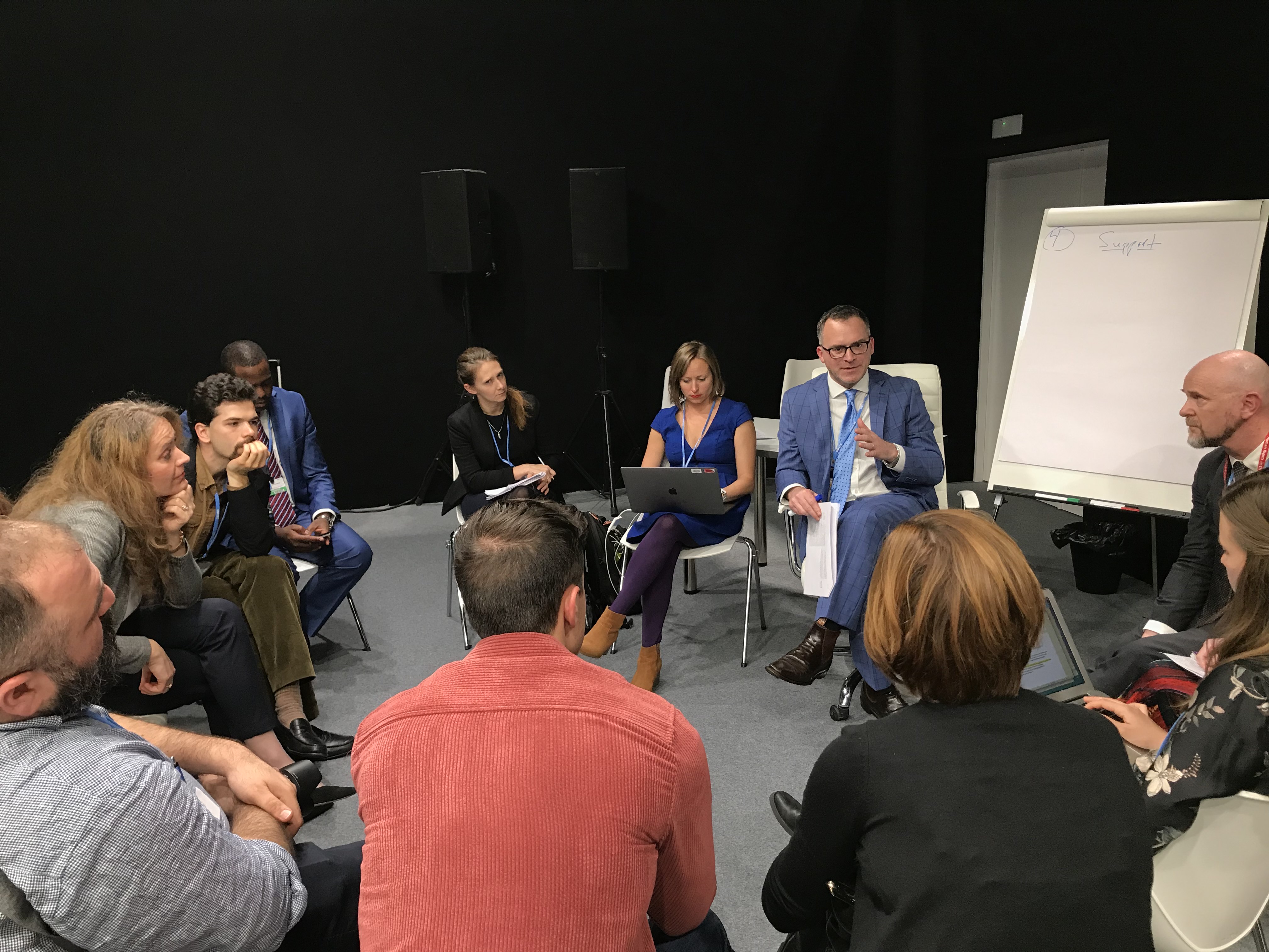 Group of people sitting in a circle, with an easel and paper next to the group.