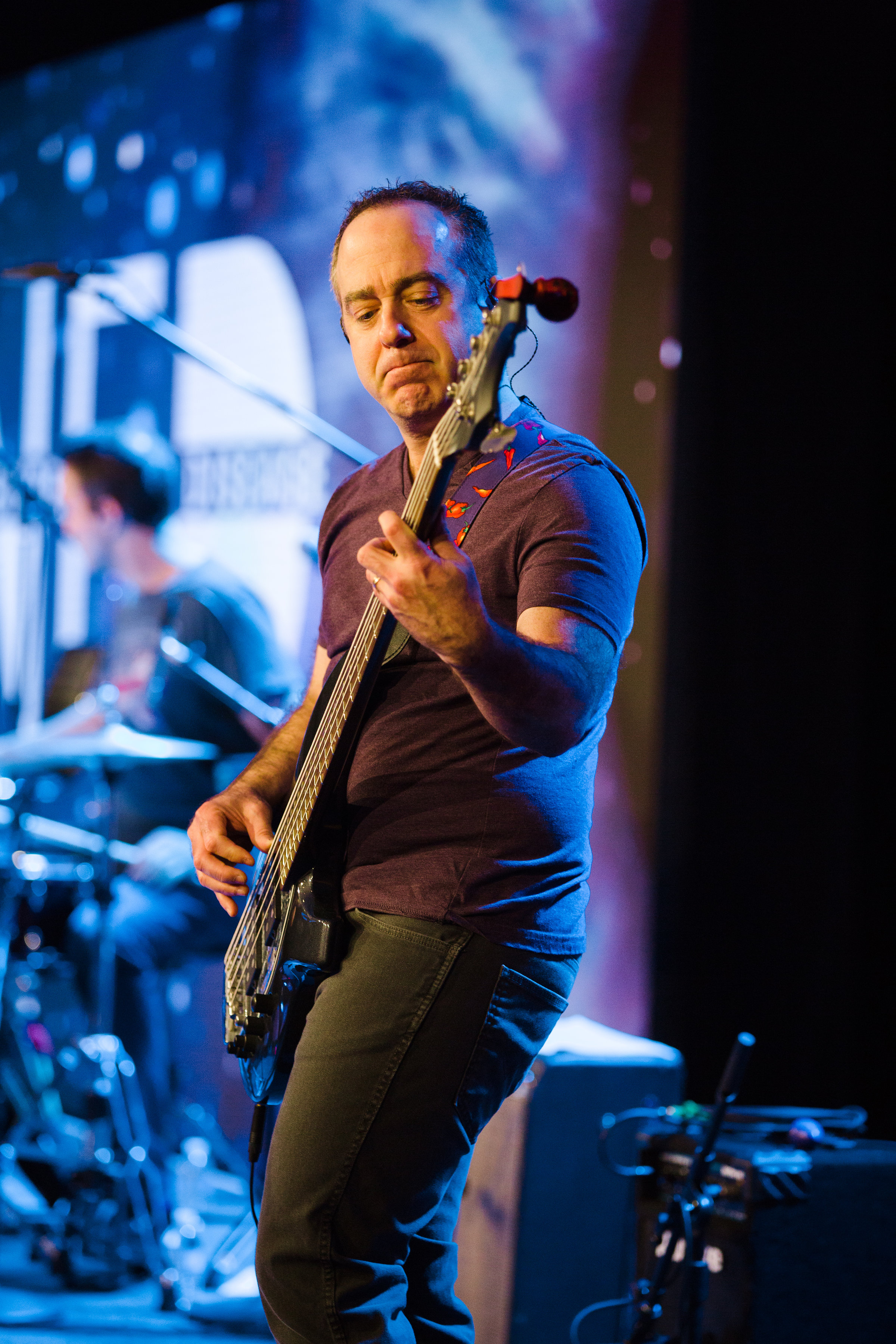 A person on a stage playing a bass guitar, with a drummer blurry in the background. 