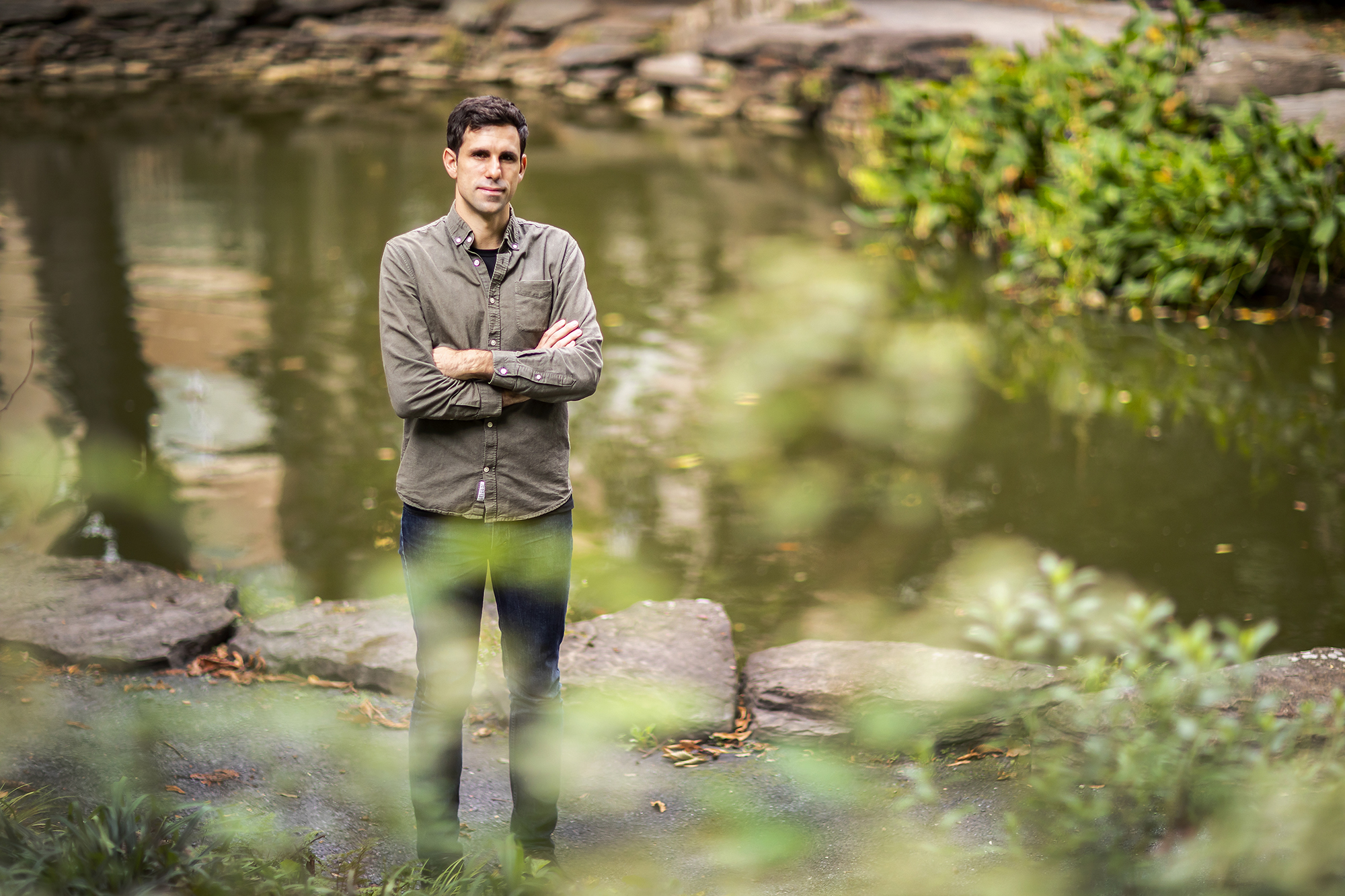 A person standing, arms crossed, in front of a pond and a rock wall around the pond's perimeter.