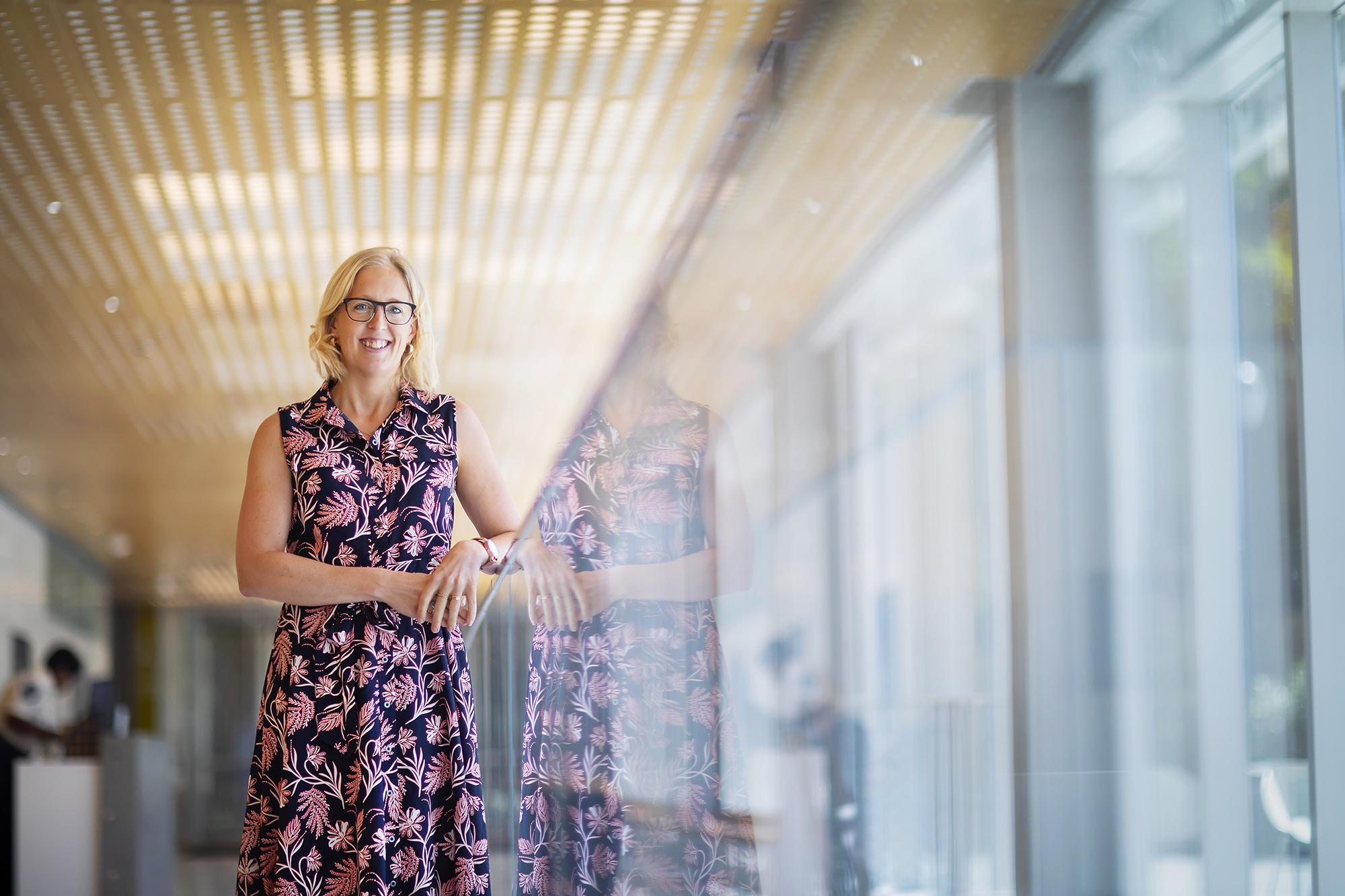 A person standing along a glass wall in a building with a yelllow waffle ceiling. 