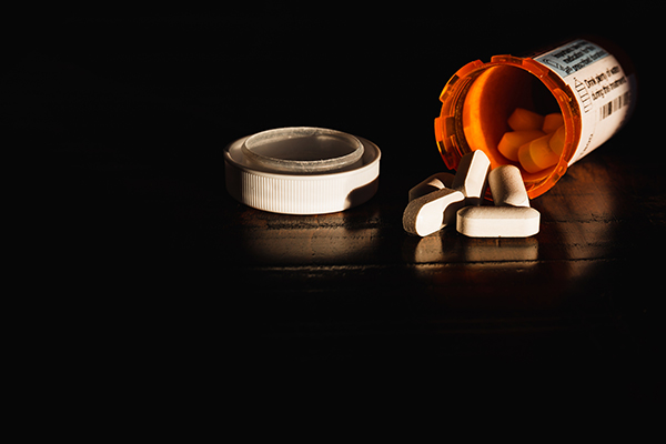 Open, overturned prescription drug bottle with pills spilling out on dark table.
