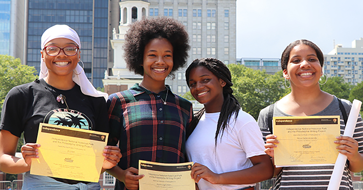 Four students hold certificates from Philadelphia Writing Project smiling and standing outdoors