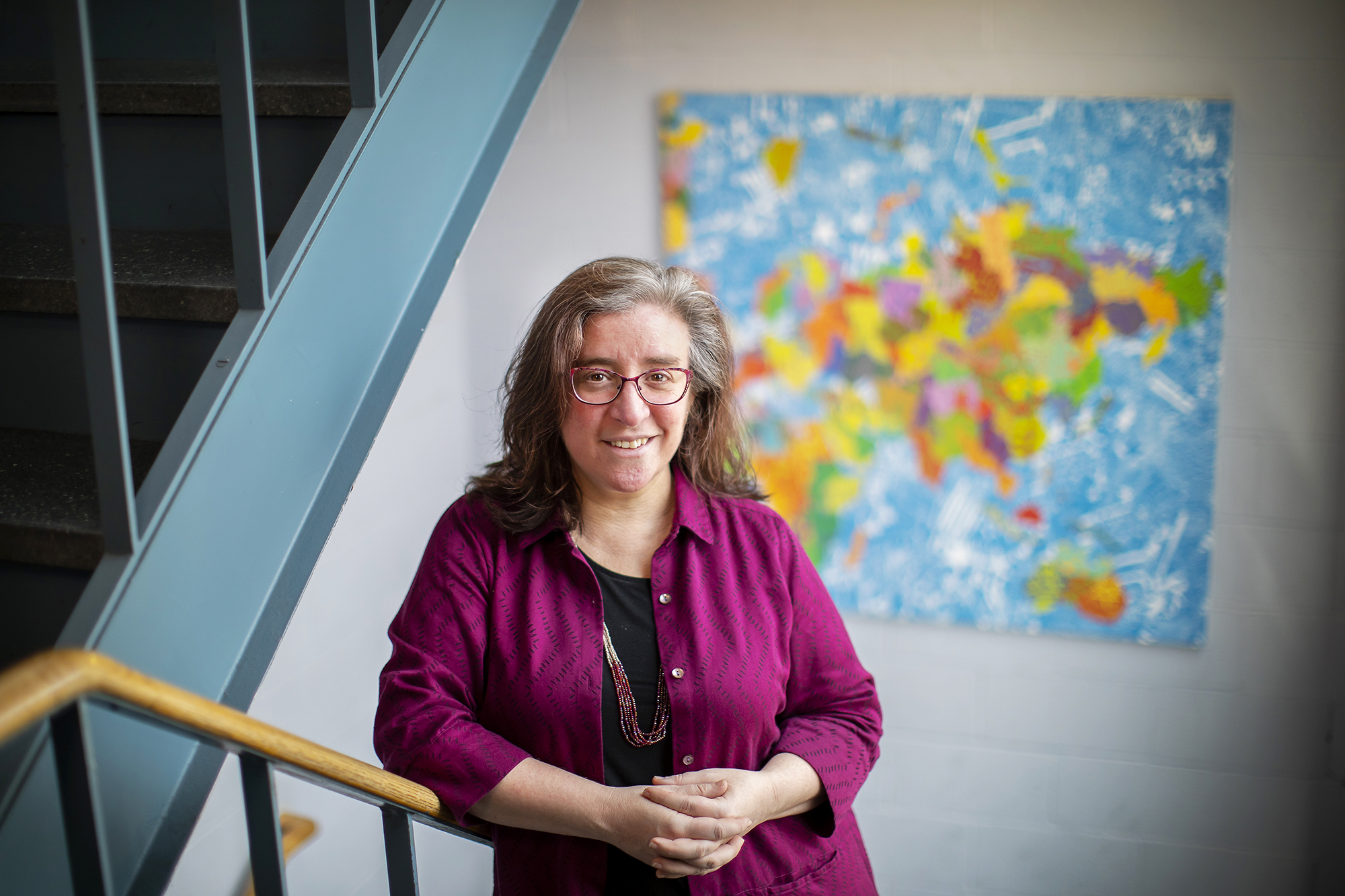 woman with clasped hands stands in a stairwell with colorful art in the background