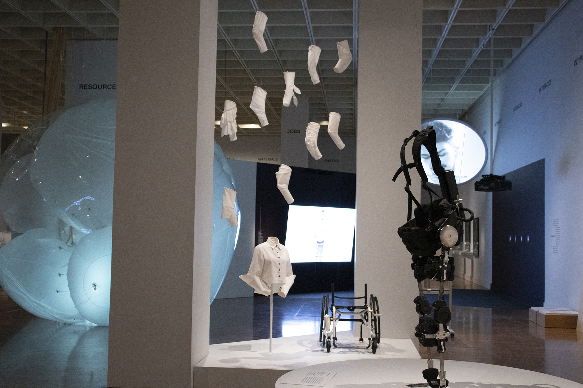 View of white garments hanging at the Designs for Different Futures exhibit, with a white shirt on a stand and a futuristic wheelchair and mechanical upright walking device on display.