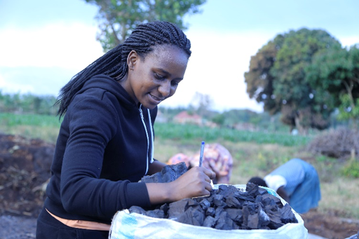 Penn alumna Catherine Nabukalu examines a bag of charcoal as two people work in a field in the background