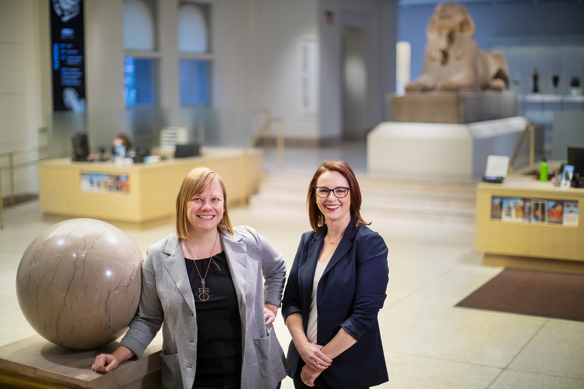 Two people standing next to a marble staircase, with stands and a sphinx blurry in the background.