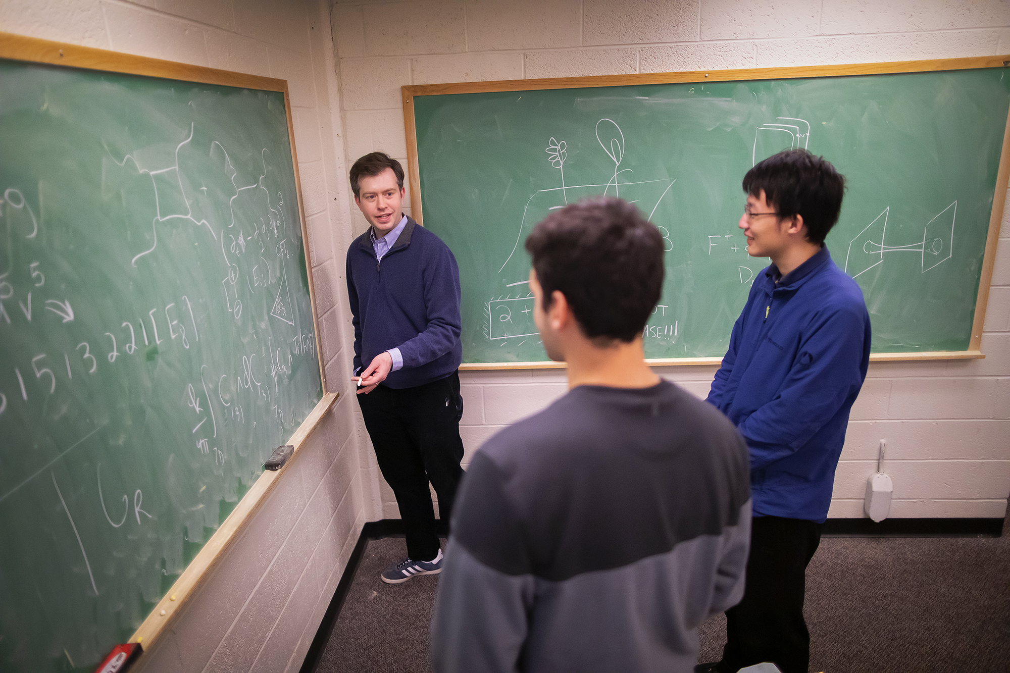 three people standing in the center of two chalkboards with one person standing at the board covered in equations with a piece of chalk in their hand