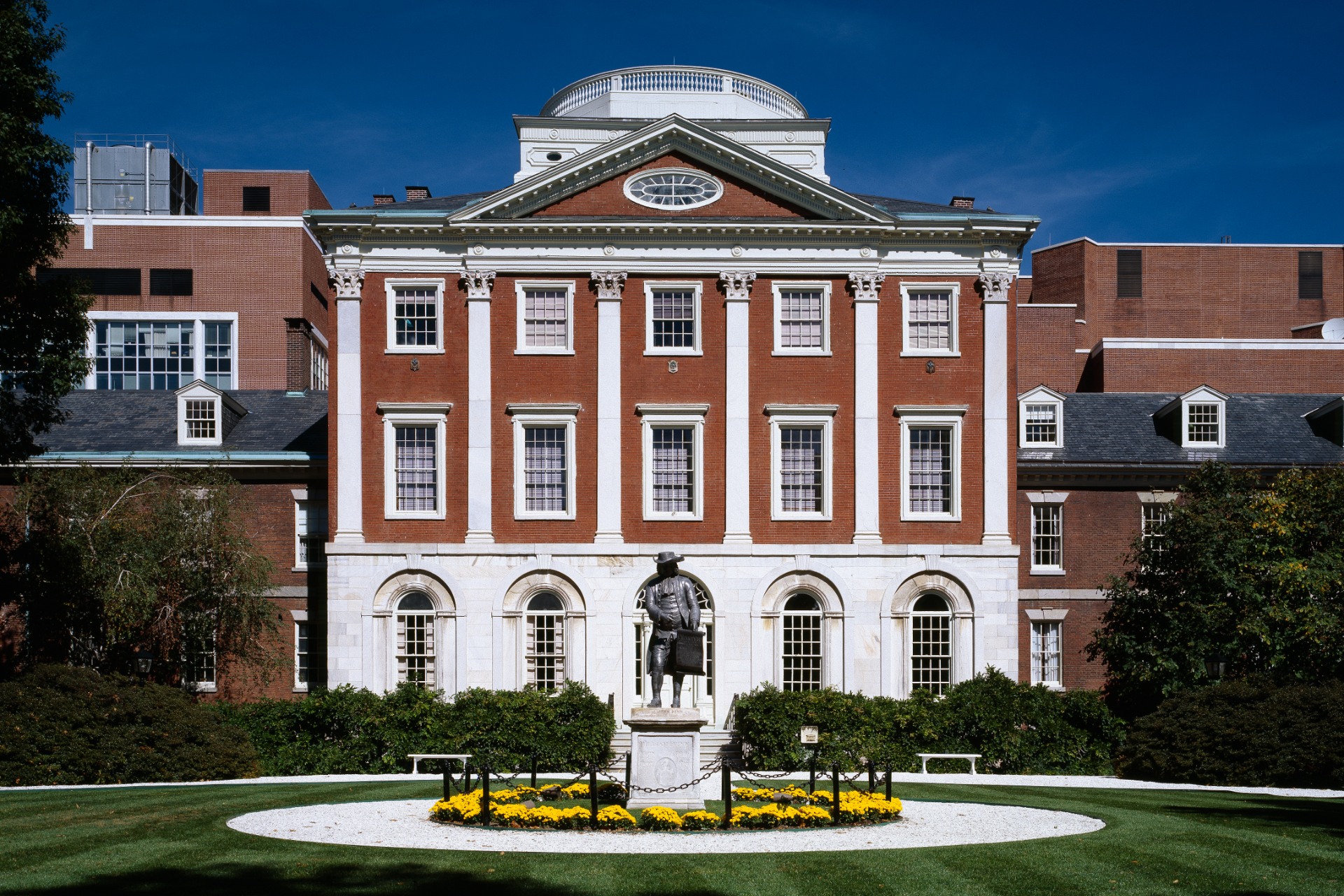 a brick building with white detailing and in front is a green field with a statue surrounded by yellow flowers