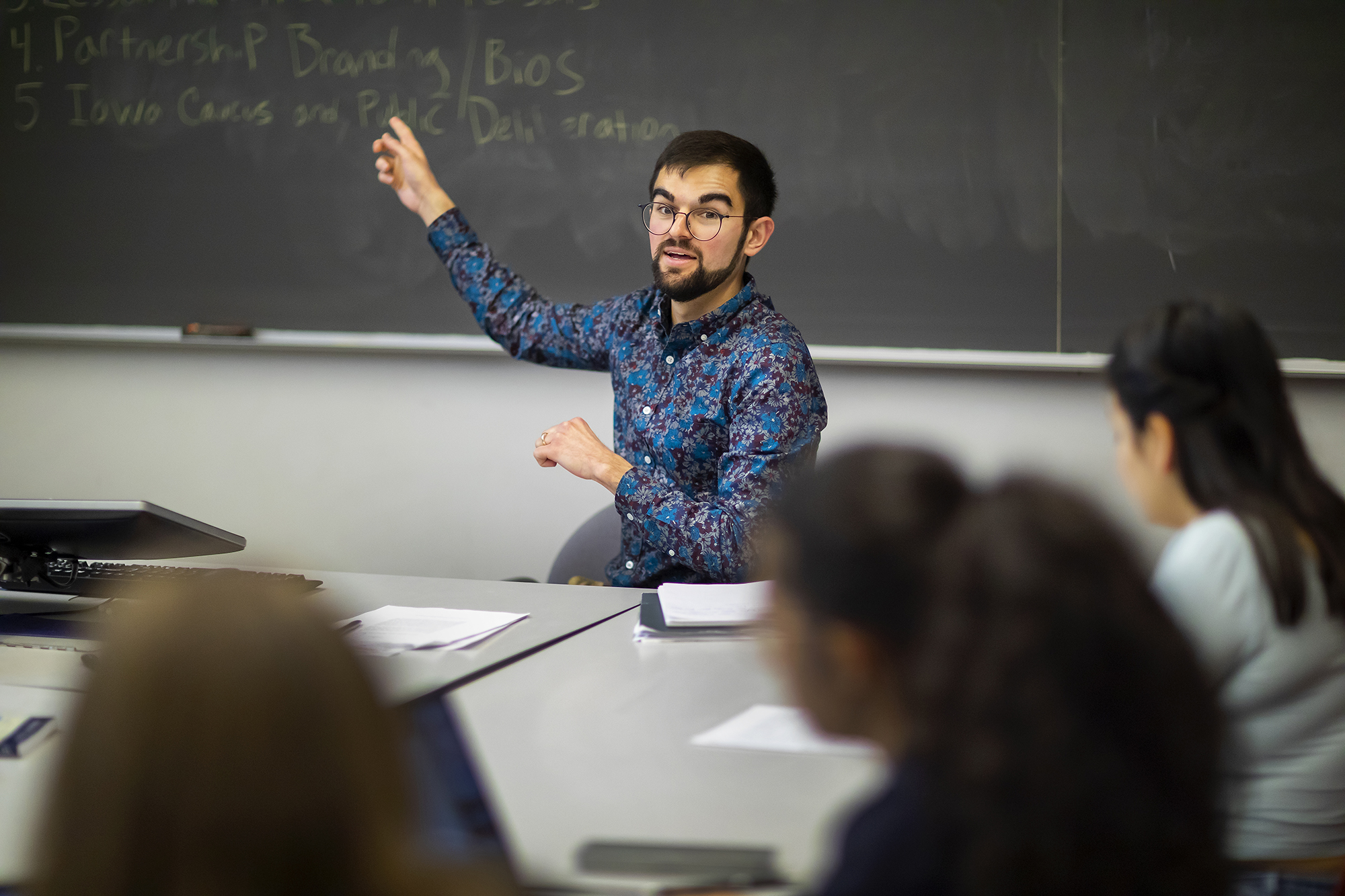 Man in flowered shirt points at a blackboard where class assignments are written