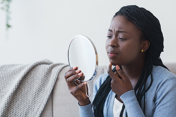 A Black person examines their skin for acne in a hand mirror.
