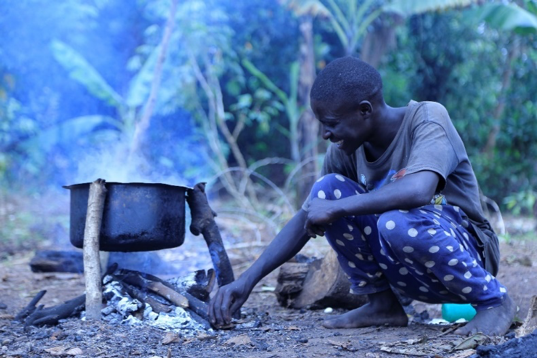 Person crouches near a cooking pot using wood to build a fire