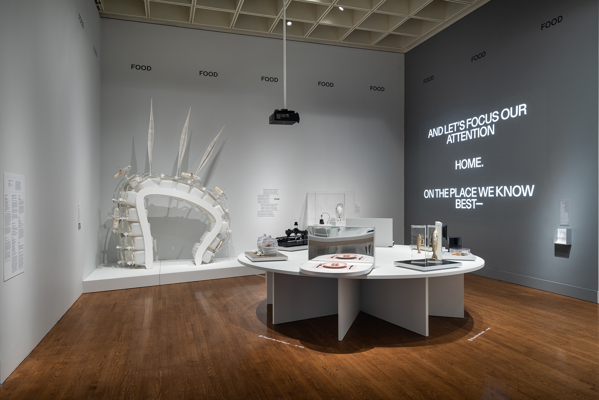 Exhibit at Designs for Different Futures, a white table with futuristic table settings, a white structure, on the wall is the text "And let's focus our attention—Home—On the place we know best.”