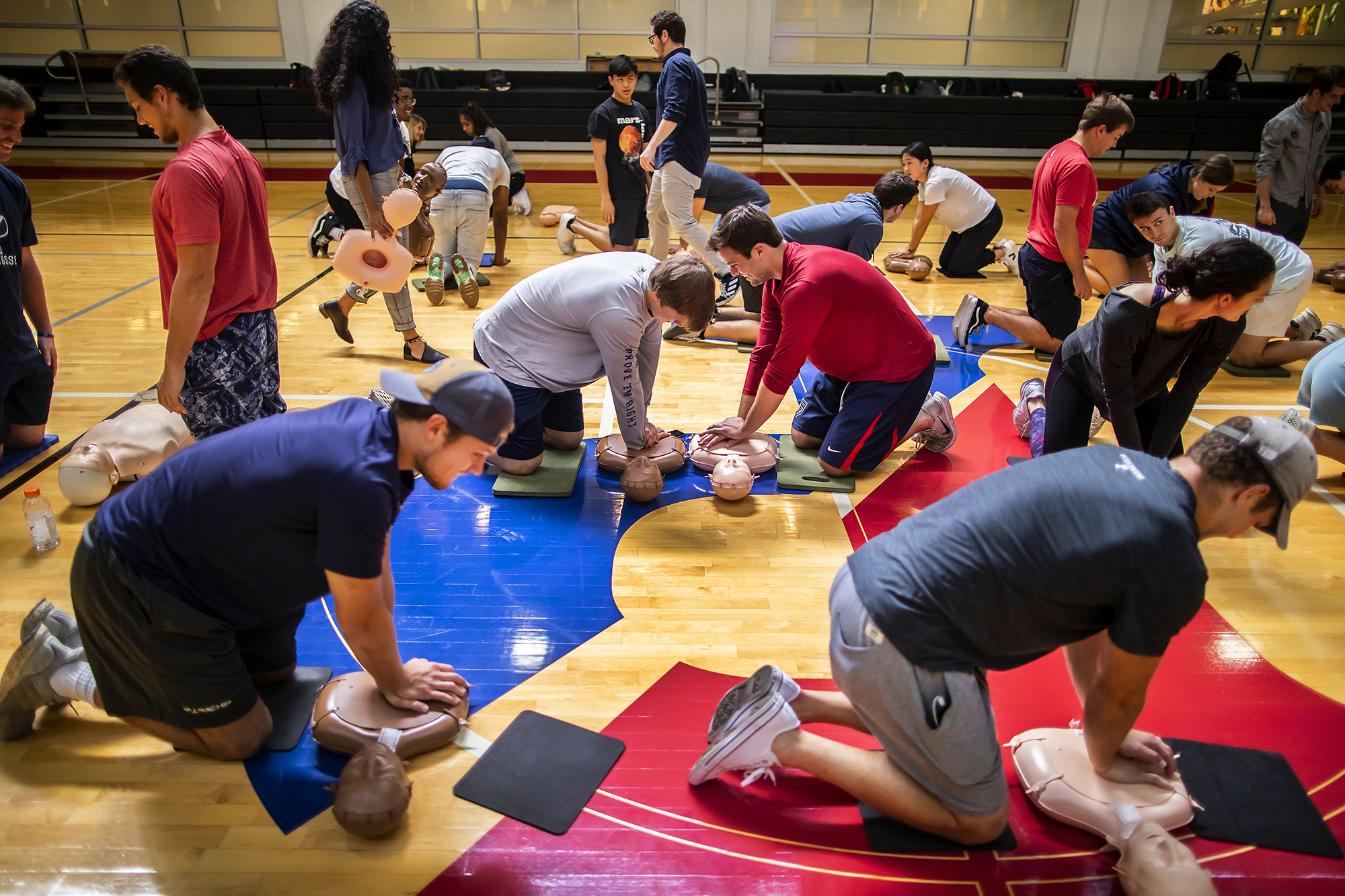 Students performing CPR on mannequins on a gymnasium floor.