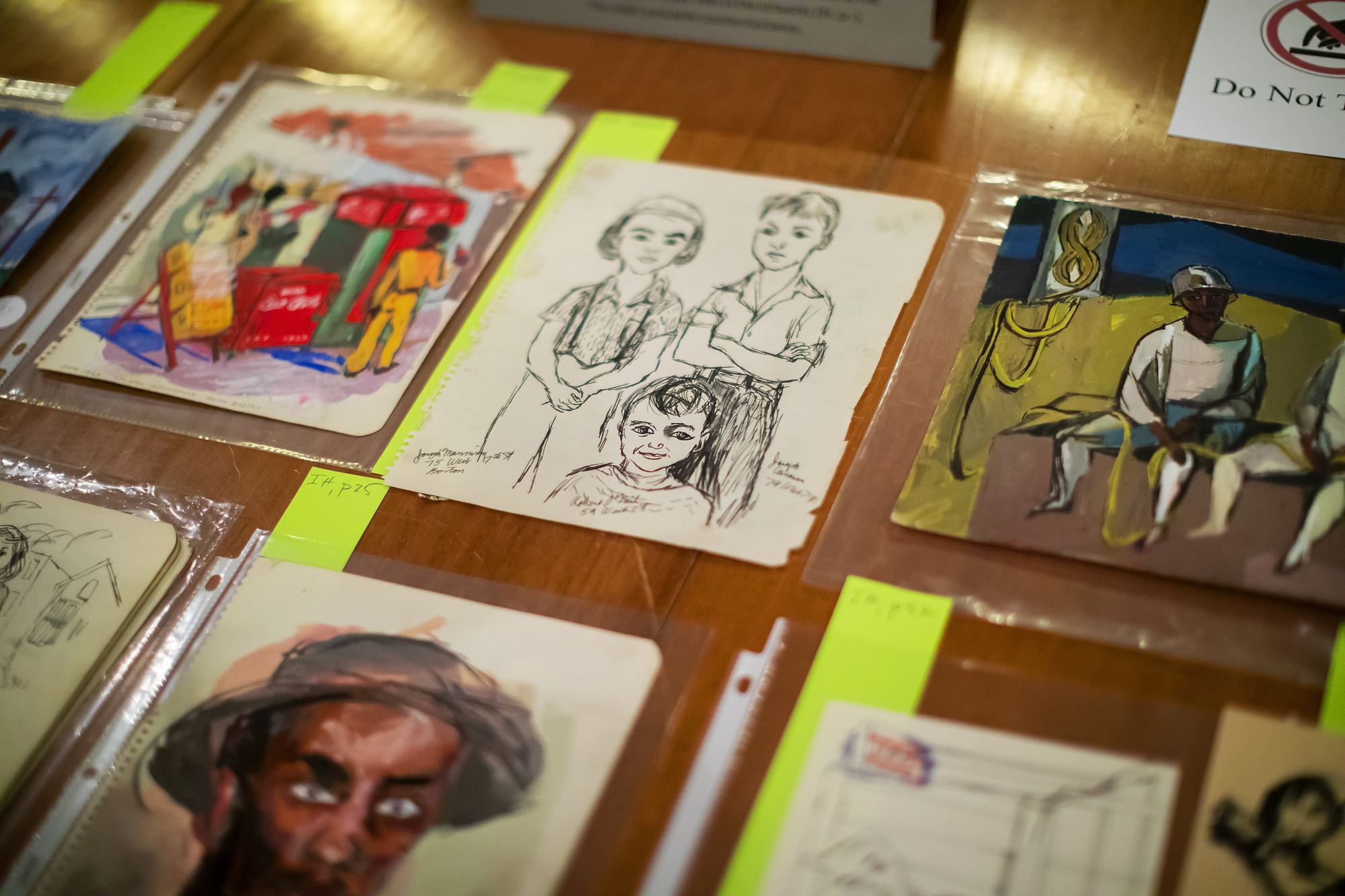 several drawings and paintings by Ashley Bryan laid out on a table under protective sheets of plastic for library cataloging. 