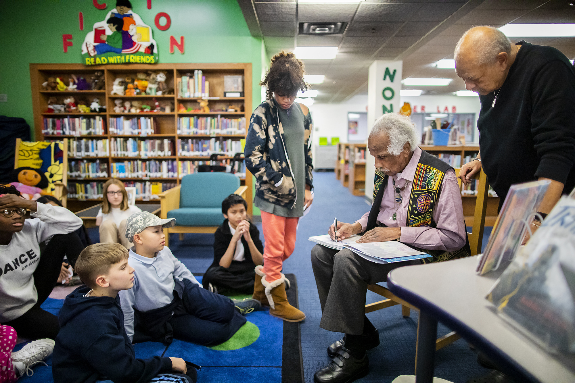 Ashley Bryan signs a copy of his book in the library at Penn Alexander School with students seated on the ground in front of him.