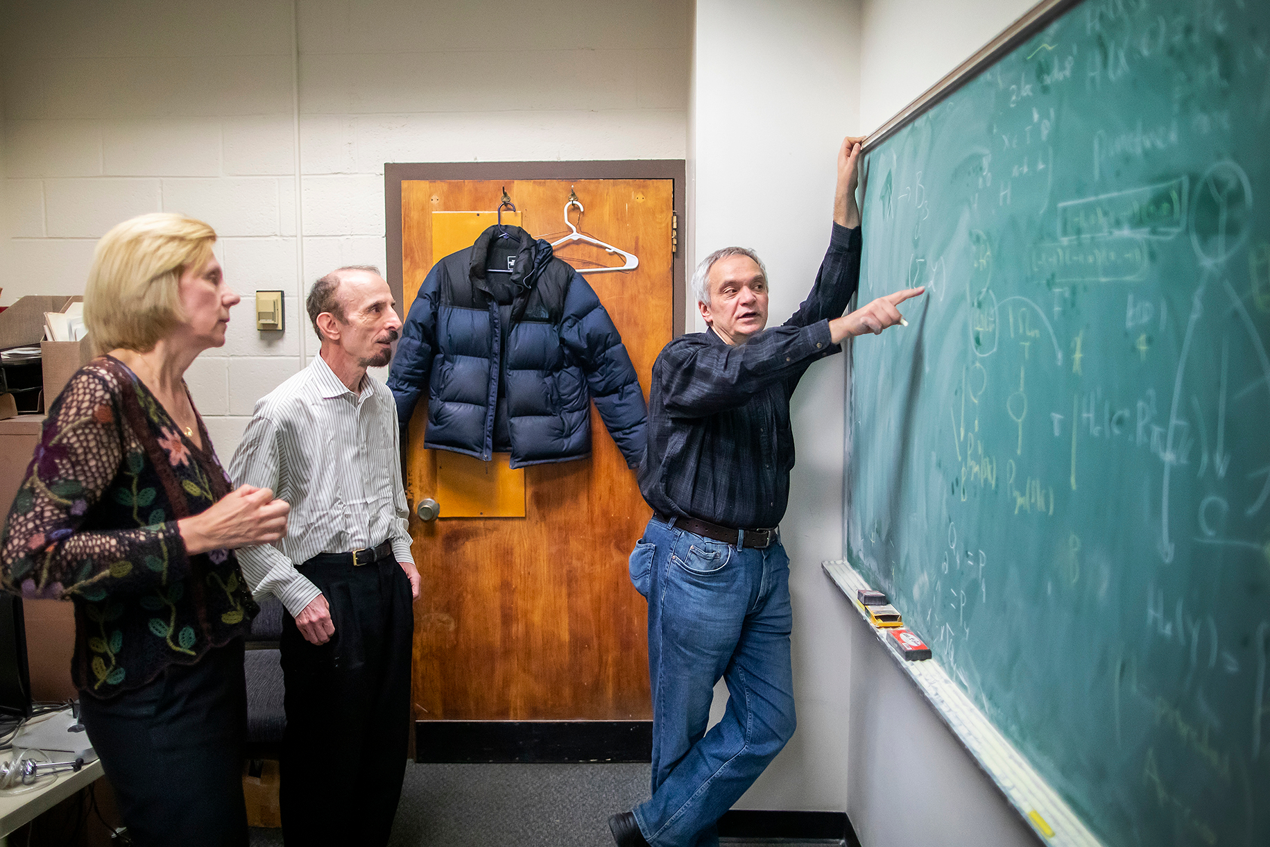 three people at a chalkboard with the person closest to the board pointing at an equation