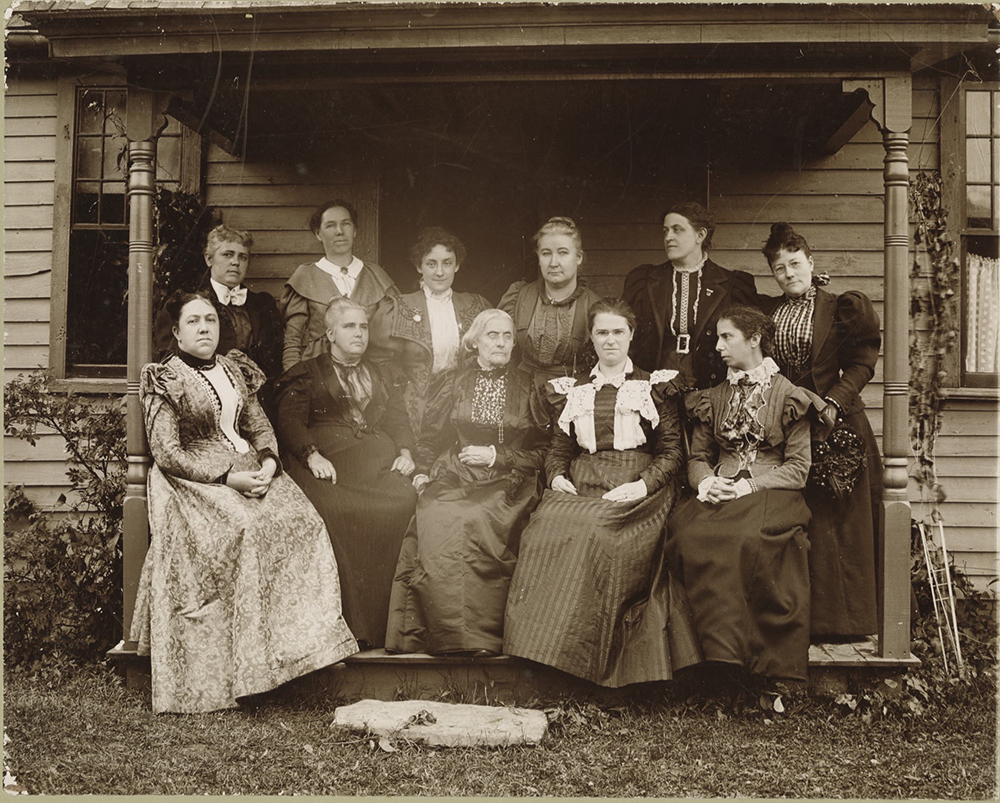 11 women from the women's suffrage women pose on a front porch, susan b. anthony seated in the center.