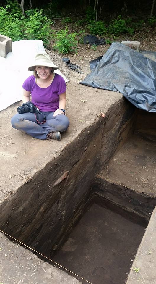 A person sitting outside next to a large whole in the dirt, part of an archaeological site.