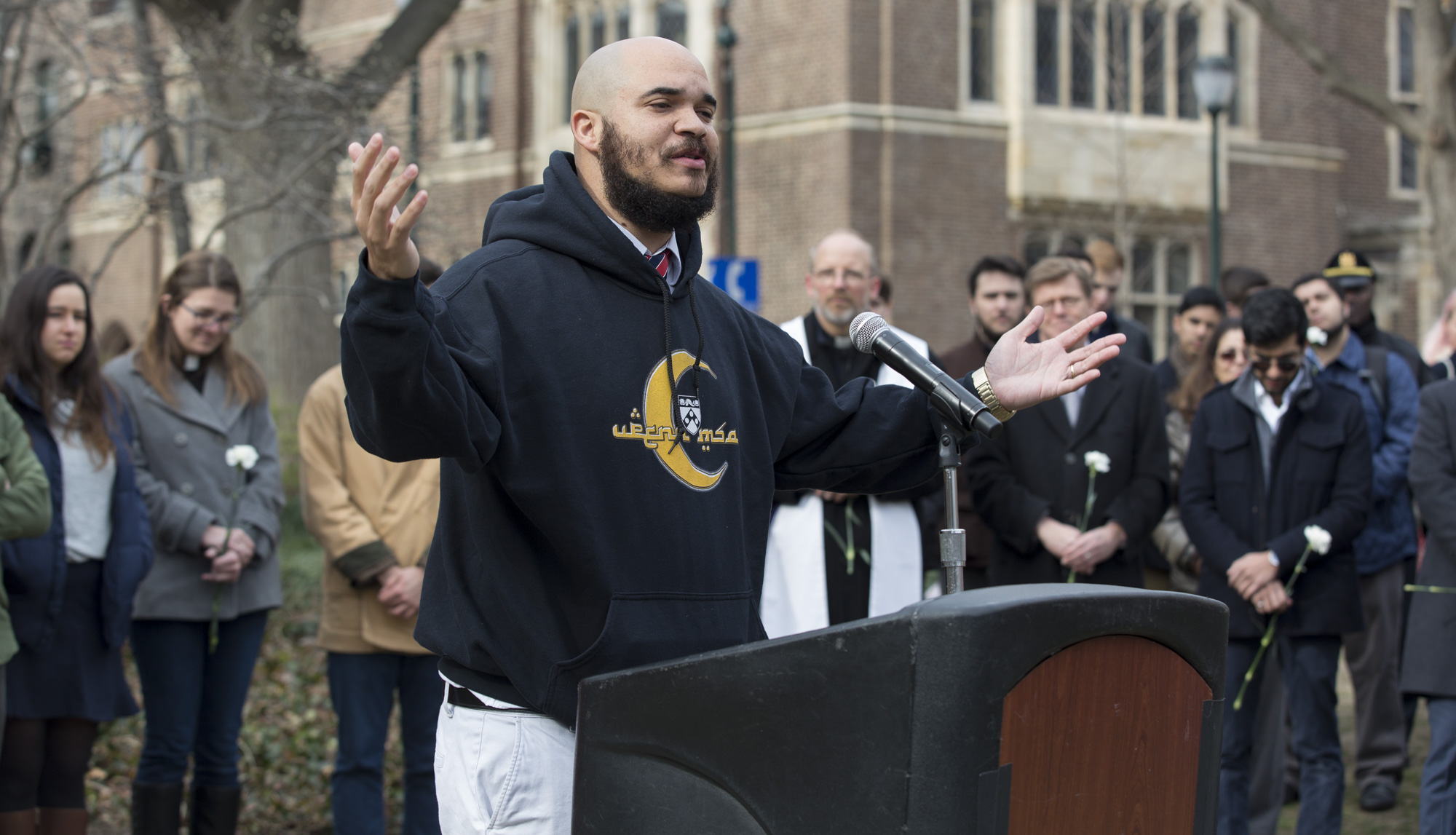 University minister standing outside at podium with a microphone, his eyes closed and arms outstretched with several people gathered behind him.