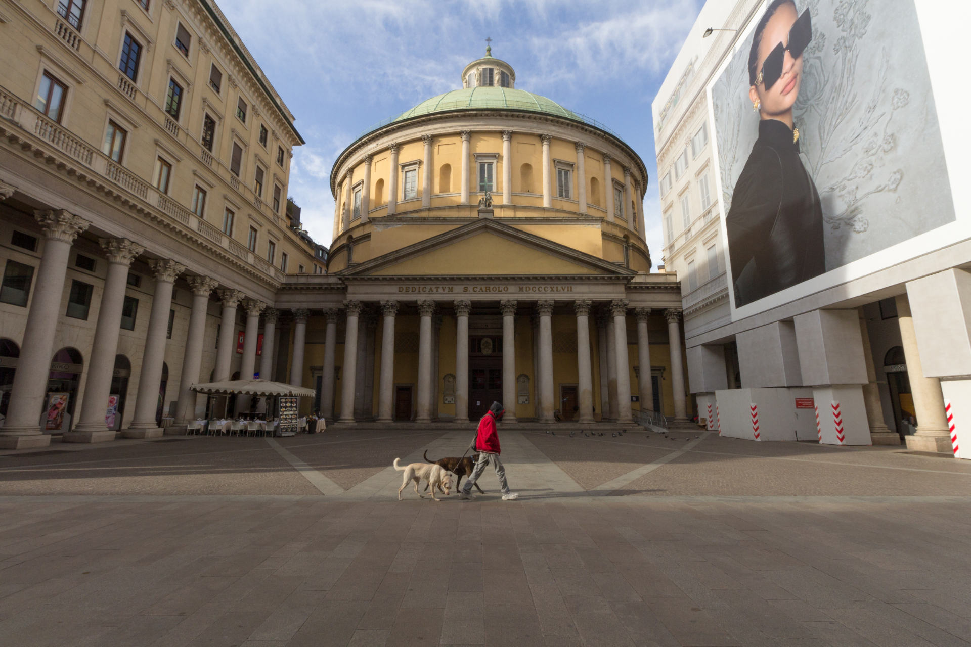 A person with two dogs walking in front of an empty plaza in front of the Cathedral of San Carlo al Corso in Milan, Italy