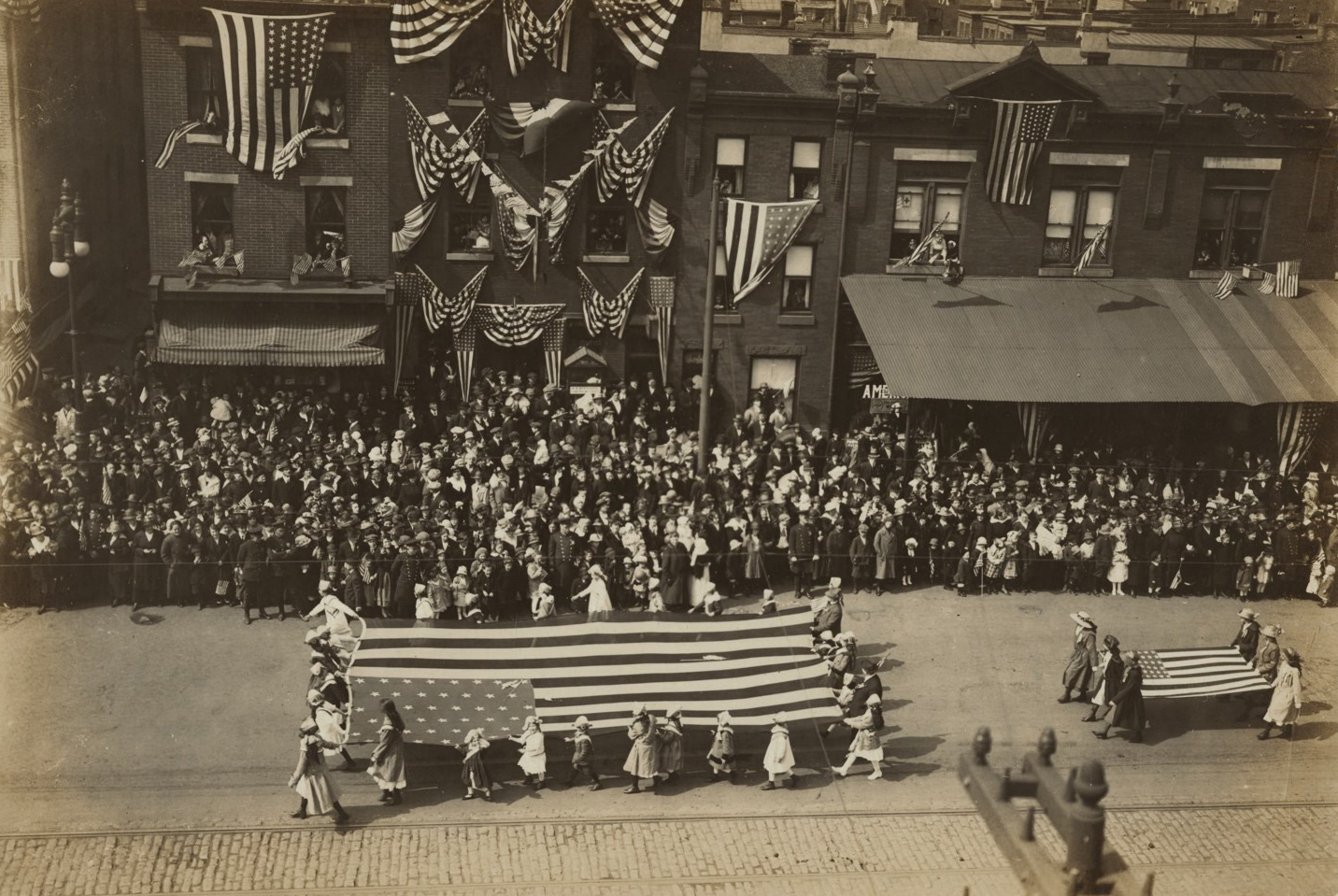 Parade from 1918 in the streets with people carrying a large American flag