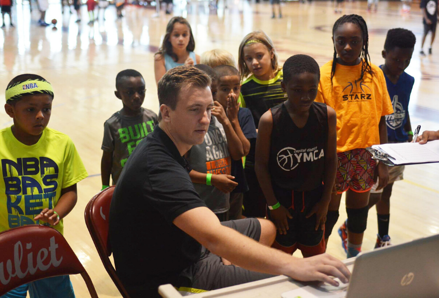 a person looking at a laptop surrounded by kids watching the screen standing in a basketball court