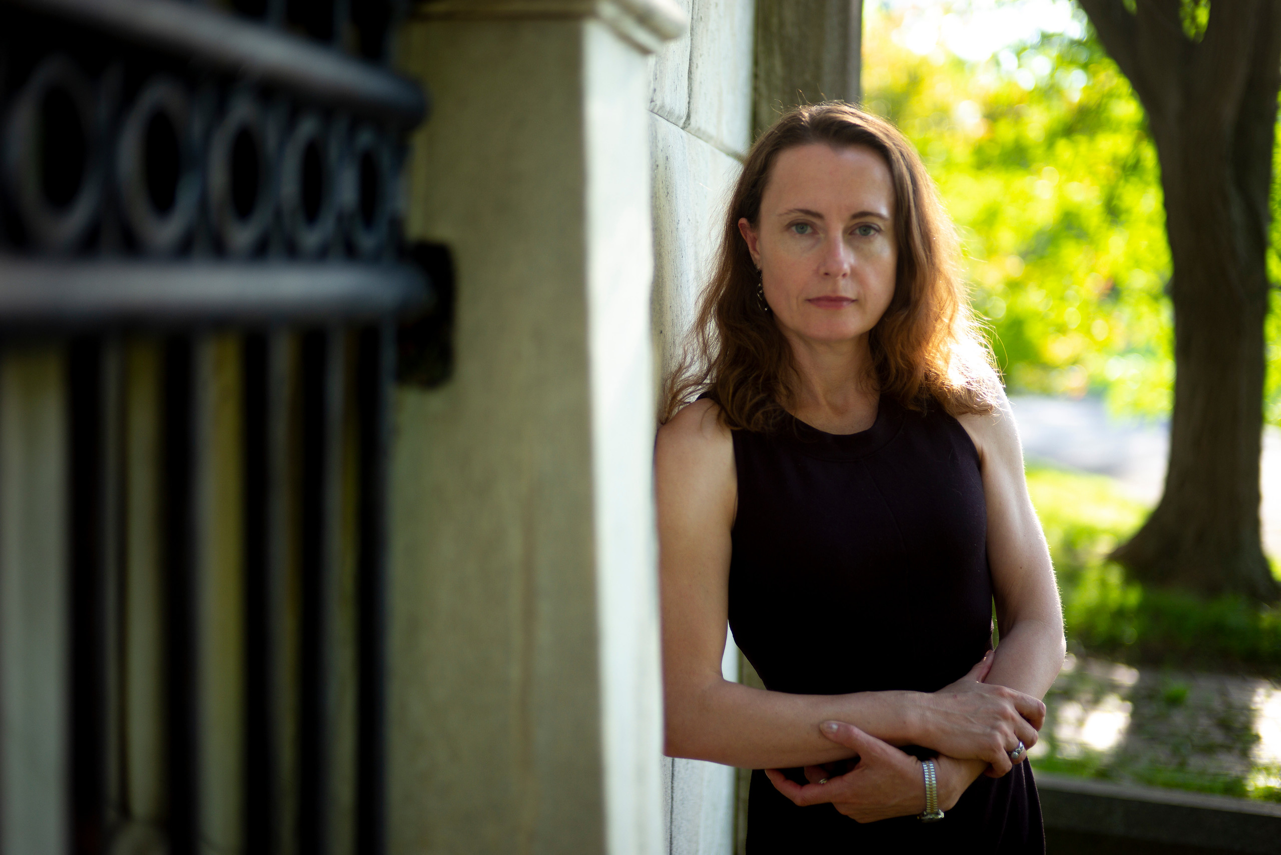 Professor stands outside leaning against a stone wall with trees in the background.