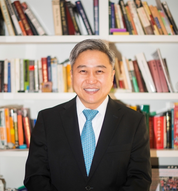 Person wearing suit and tie stands in front of book case, smiling at camera