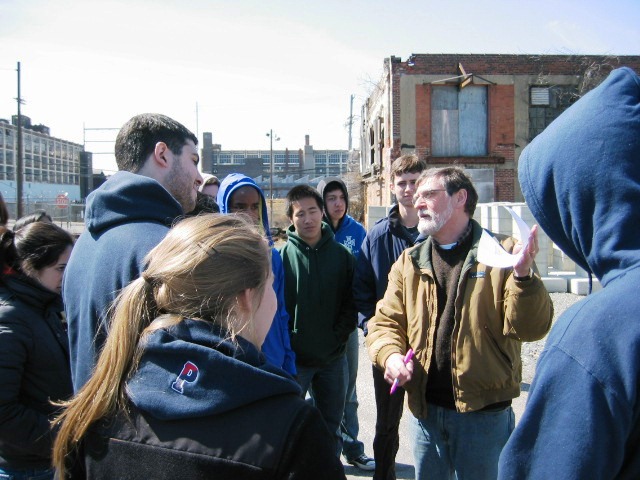 Person with beard and glasses speaks to group of college-age students in front of old industrial buildings.