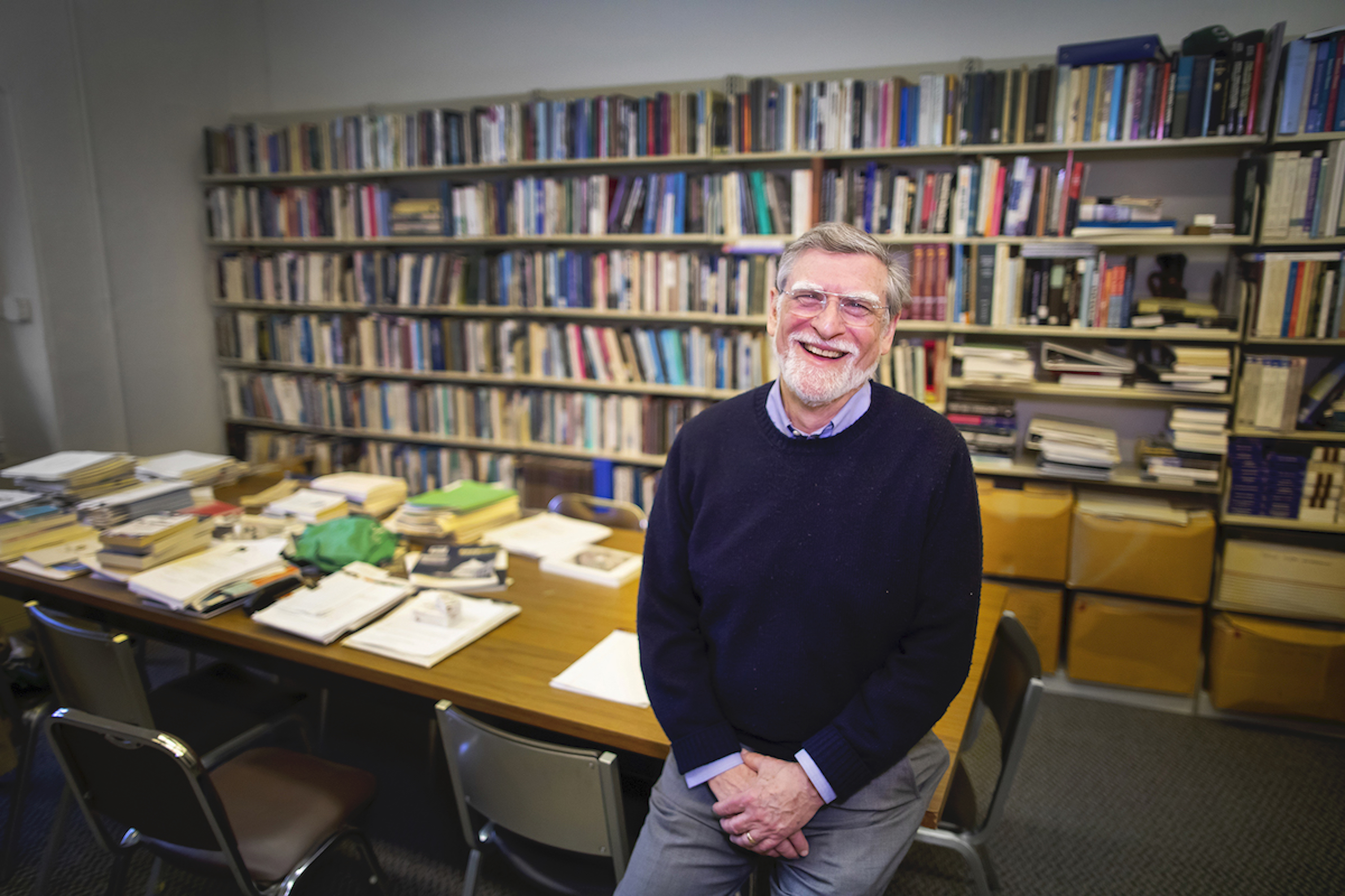 Person sits on corner of conference table with wall of books behind them, smiling at the camera.