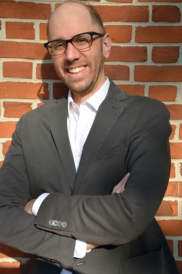 Person wearing glasses leans against red brick wall with arms folded, smiling a