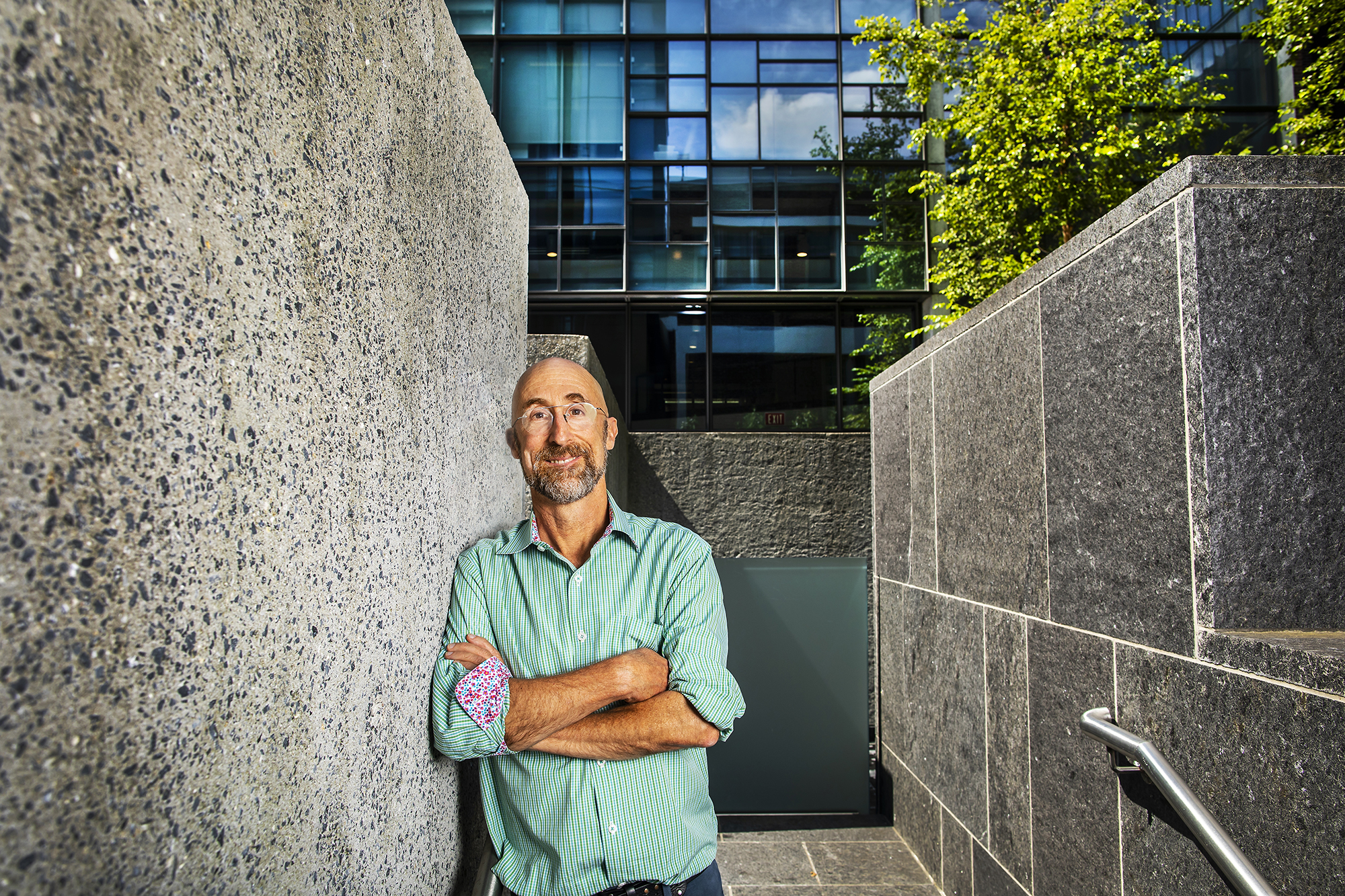 A person with arms crossed at the chest standing outside between two rock walls, in front of a glass building.